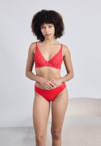 Rdeča čipkasta modrček in ujemajoč komplet hlačk z cvetličnimi vzorci, valovitimi robovi in majhnim lokom na pasu. Gladka tekstura.