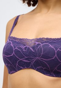 Mežģīņu bralete dziļi purpursarkanā krāsā ar ziedu rakstu, ar viļņotu malu un regulējamiem siksniņām. Mīksta tekstūra un izliekti kausi.