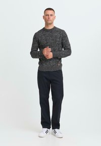 Man staat met zijn handen gekruist, draagt een donkergrijze gebreide trui, zwarte broek en witte sneakers met zwarte strepen.