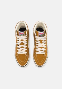 Brązowe, zamszowe sneakersy za kostkę z białymi sznurówkami, wyraźnym przeszyciem i czarnymi akcentami, z logo Vans na języku i boku.