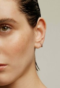 Aro de plata con acentos puntiagudos, llevado en la oreja izquierda. Acabado metálico suave que complementa el tono de piel. Primer plano de las características faciales.