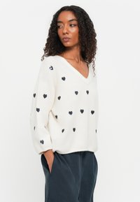 Maglione bianco, morbido e oversize, con scollo a V e motivo di cuori in paillettes blu navy. Tessuto morbido, maniche lunghe e tasche laterali. Design casual.