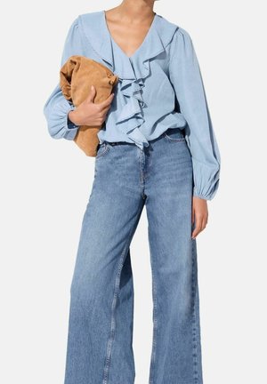 Person iført lyseblå flæsebluse og vidde jeans holder en tan foldet ruskindsjakke mod en ensfarvet baggrund.