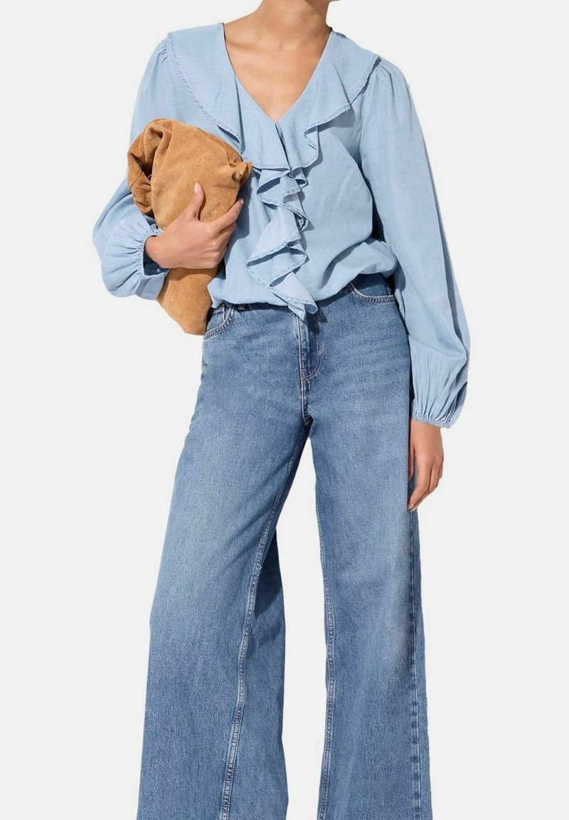 Personne portant un chemisier bleu clair à volants et un jean à jambes larges tenant une veste en daim beige pliée sur un fond uni.