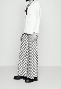 Blazer bianco sopra un top nero, abbinato a pantaloni con ampi quadri bianchi e neri e scarpe nere spesse. Tessuto morbido, vestibilità rilassata, design minimalista.