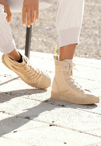 Zapatillas beige de caña alta con un upper de lona texturizada, puntera redonda y suela de goma gruesa, combinadas con unas zapatillas de perfil bajo en el mismo color.