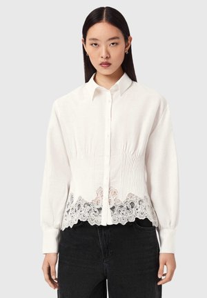 Biała koszula na guziki z długimi rękawami, z marszczonym przodem i ozdobną koronką na dole. W połączeniu z ciemnymi dżinsami.