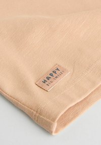 Barack színű szövet, amely puha tapintású. Egy varrott címke található rajta, amelyen vastag betűkkel olvasható a "HAPPY" felirat egy téglalap alakú háttéren.