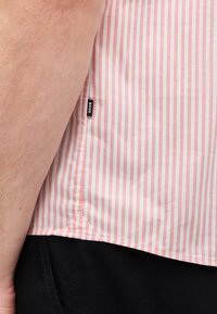 Κοντινό πλάνο από το χέρι ενός ατόμου δίπλα σε πουκάμισο με ροζ και λευκές κάθετες ρίγες και ετικέτα «BOSS», συνδυασμένο με μαύρο παντελόνι.