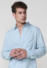 Camicetta azzurra chiara con bottoni, realizzata in tessuto morbido, con maniche lunghe, colletto classico e taschino sul petto con un ricamo discreto.