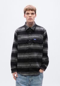 Πουκάμισο από φανέλα με μαύρες και γκρι ρίγες, γιακά, κουμπιά στο μπροστινό μέρος και δύο τσέπες στο στήθος.  Διαθέτει μικρό μπλε λογότυπο στην αριστερή τσέπη.