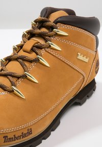 Brązowe i beżowe skórzane buty turystyczne z złotymi metalowymi oczkami, brązowymi sznurówkami i wytrzymałą czarną podeszwą, z logo Timberland.