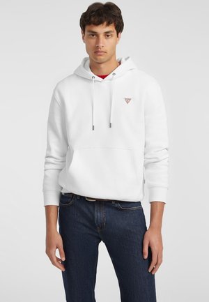 Sudadera blanca con cordones ajustables, bolsillo frontal y pequeño logo triangular; combinada con jeans azul oscuro. Tejido suave, ajuste relajado.