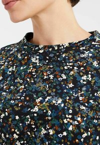 Donna che indossa una maglietta scura con motivo floreale, decorata con piccoli fiori bianchi, blu, verdi e color ruggine, e con scollatura rotonda.