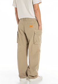 Pantalones cargo beige con un corte relajado, que cuentan con una cintura elástica, bolsillos laterales y una etiqueta de marca naranja en la parte trasera.