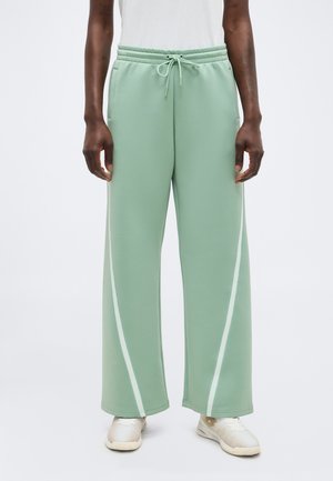 Osoba nosząca miętowe szerokie spodnie z białymi paskami po bokach, białe trampki i prostą białą bluzkę, stojąca na jednolitym tle.