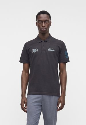 Homme portant un polo noir avec des empiècements bleus sur la poitrine et la manche, associé à un pantalon gris clair, debout devant un fond blanc.