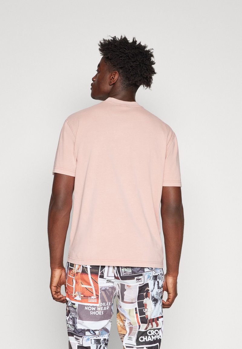 T-shirt rose pâle à manches courtes et col rond. Le tissu semble lisse. Associé à un short à motifs présentant diverses impressions graphiques.