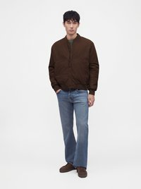 Jeune homme portant une veste marron zippée, un jean bleu et des chaussures marron, debout avec une main dans la poche sur fond blanc uni.