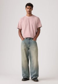 Svetloružové bavlnené tričko s krátkymi rukávmi, doplnené širokými džínsami modrého tieňovaného denimu. Oblečenie dopĺňajú tmavé topánky.