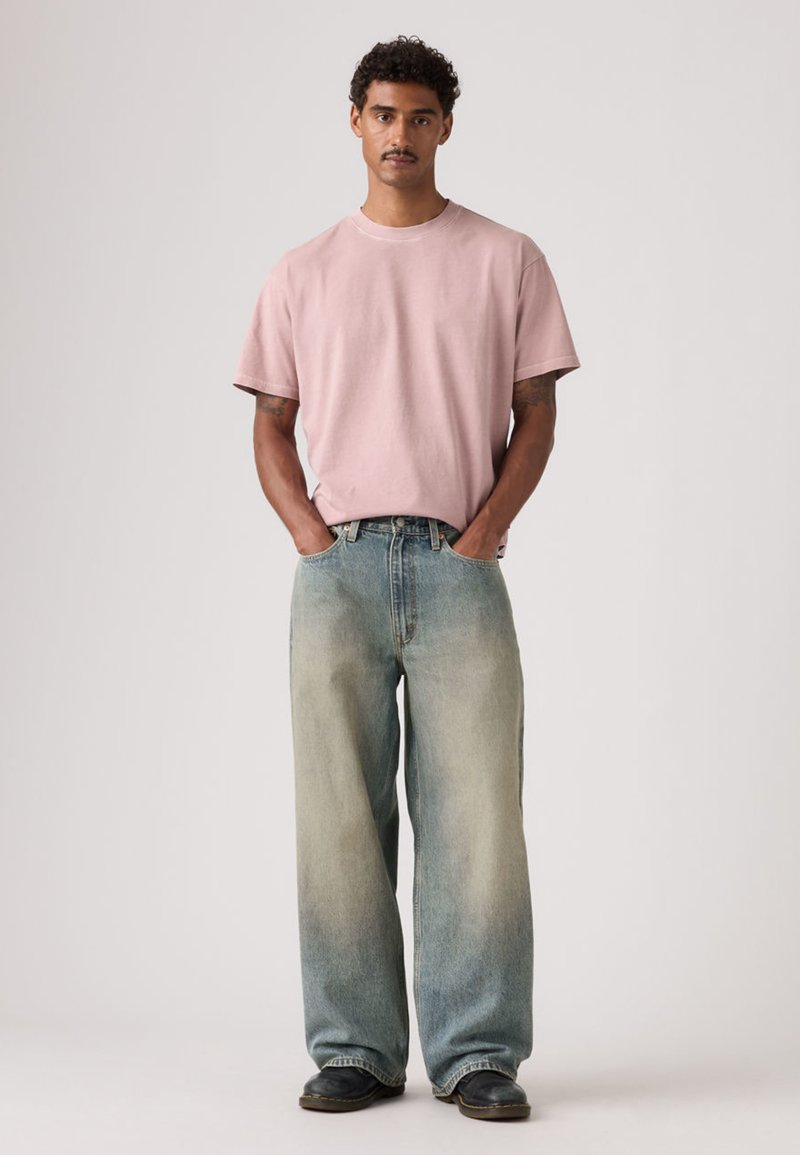 Svetloružové bavlnené tričko s krátkymi rukávmi, doplnené širokými džínsami modrého tieňovaného denimu. Oblečenie dopĺňajú tmavé topánky.