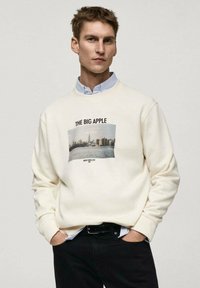 Kremna športna majica z dolgimi rokavi, ki prikazuje grafiko newyorške panorame in besedilo "VELIKA JABOČKA" zgoraj. Ima okroglo vratno odprtino.