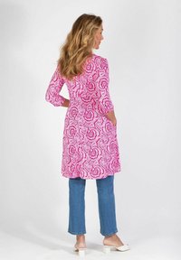 Długa różowa sukienka w wzory z trzema ćwierć rękawami, z nadrukiem paisley, w połączeniu z niebieskimi dżinsami i białymi flatami.