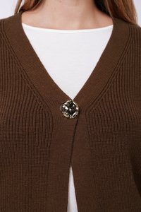 Καφέ πλεκτή ζακέτα με βαθύ V-λαιμόκοψη και ένα γυαλιστερό, διακοσμητικό κουμπί στο κλείσιμο, φορεμένη πάνω από λευκό πουκάμισο.