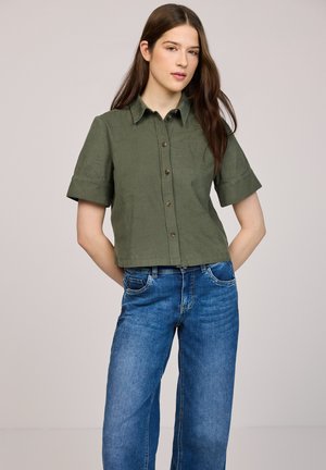 Nuori nainen, jolla on pitkät ruskeat hiukset ja joka käyttää oliivinvihreää lyhythihamaista napitettavaa paitaa sekä sinisiä farkkuja, seisoo kädet selän takana.