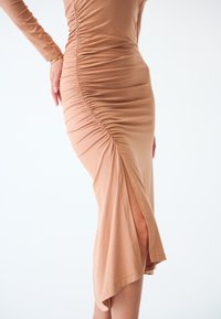 Abito lungo aderente in tessuto beige chiaro con arricciature laterali e uno spacco alto fino alla coscia, dotato di maniche lunghe e una texture liscia e leggermente elastica.