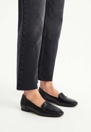 Black leather loafers with a flat heel feature a stitched strap detail and a smooth finish. Worn with dark denim pants that have frayed hems.