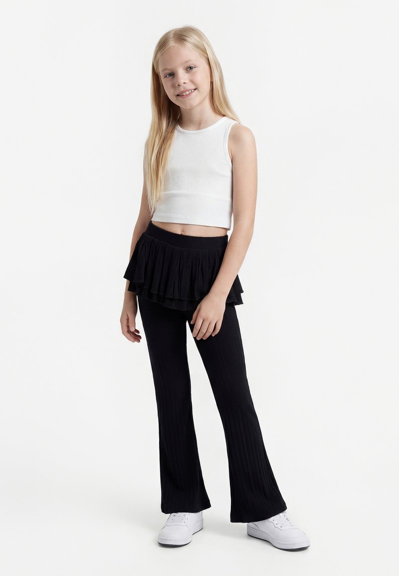 Jeune fille aux longs cheveux blonds portant un crop top blanc sans manches, une jupe-pantalon noire superposée et des baskets blanches, debout et souriante.