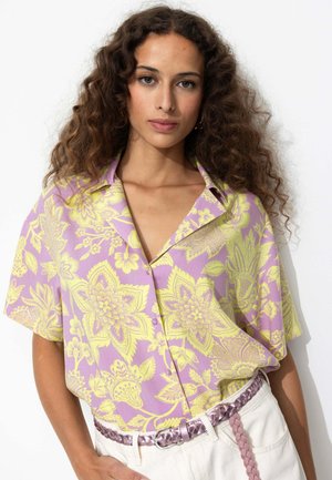 Chemise à manches courtes de couleur violette avec un motif floral jaune, dotée d'un col et de boutons. Portée avec une ceinture tressée de couleur claire.