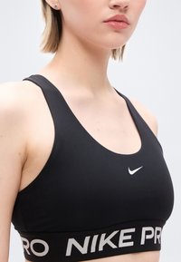 Κοντινό πλάνο ενός ατόμου που φοράει μαύρο αθλητικό σουτιέν Nike Pro με λευκό λογότυπο swoosh και ελαστική ταινία με το εμπορικό σήμα.