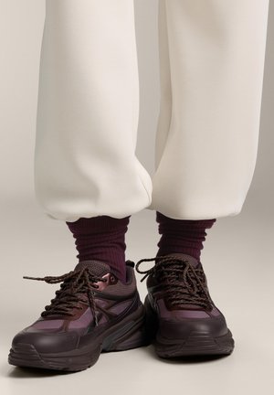 Persoană poartă adidași mov închis cu șosete bordo și pantaloni de culoare alb murdar strânși la glezne, stând pe un fundal neutru.