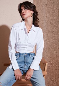 Camicia bianca a maniche lunghe con bottoni, corta, con colletto strutturato e design slim, abbinata a jeans a vita alta blu chiaro.