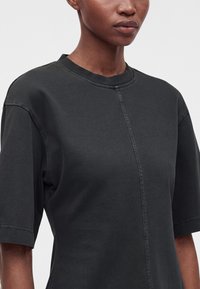 Gros plan d'une personne portant un t-shirt noir uni à col rond avec des coutures visibles au centre et sur les manches.