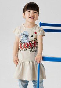 Beżowa sukienka z krótkim rękawem i marszczoną spódnicą, z grafiką Sticha, czerwonymi sercami i literami w wzór lamparta. Dżinsowe spodnie i niebieskie krzesło.