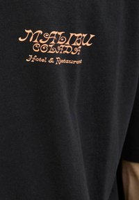 Musta puuvillapaita, jossa on oranssi teksti, joka lukee "MALIBU COLADA Hotelli & Ravintola" kursiivilla.