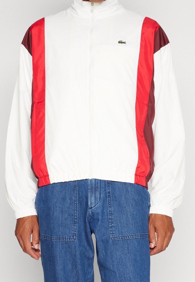 Biała kurtka zapinana na zamek z czerwonymi i bordowymi panelami bocznymi, elastycznymi mankietami i pasem. Na piersi znajduje się małe zielone logo.