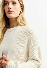Cardigan in maglia color crema con un motivo testurizzato, apertura frontale e maniche larghe. Indossato sopra una maglietta leggera, evidenziando una vestibilità morbida e materiale soffice.