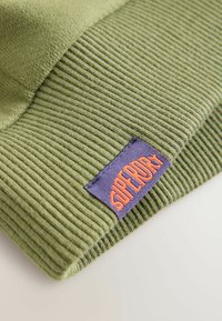 Gorro de punto verde con textura acanalada. Presenta una etiqueta morada prominente con texto naranja que dice "Superdry". Material suave y diseño casual.
