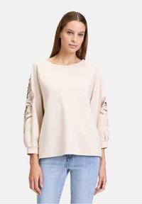 Sweat-shirt rose clair avec des manches raglan, affichant des motifs floraux complexes et des accents sur les manches. Texture douce et coupe décontractée.