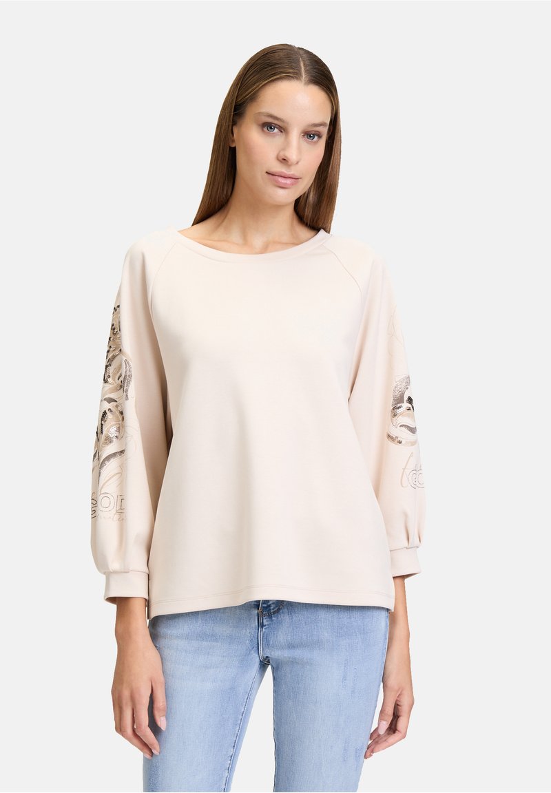 Sweat-shirt rose clair avec des manches raglan, affichant des motifs floraux complexes et des accents sur les manches. Texture douce et coupe décontractée.