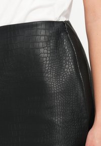 Czarna spódnica z ekoskóry o teksturowanym wzorze krokodylej skóry. Model o średnim stanie z ukrytym suwakem po bokach, prezentujący gładkie, błyszczące wykończenie.