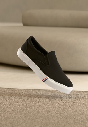 Μαύρο αθλητικό παπούτσι χωρίς κορδόνια με λευκή σόλα και λεπτομέρεια με ρίγες σε κόκκινο, λευκό και μπλε στο πλάι, αιωρούμενο πάνω από μπεζ χαλί σε ένα ουδέτερο δωμάτιο.