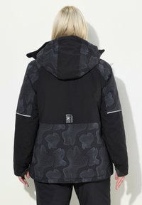 Svart isolerad jacka med mönstrad baksida med abstrakta linjer, huva och reflekterande detaljer på ärmarna.