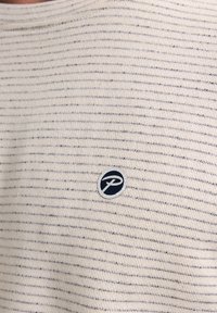 Camisa de punto en color crema claro con rayas horizontales en azul marino, que presenta un parche con un logo circular en el lado izquierdo del pecho.