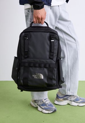 Personne tenant un sac à dos noir North Face par sa poignée supérieure, portant un jean clair et des baskets New Balance bleu gris sur un sol vert.