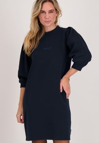 Marineblauwe sweatshirtjurk met een losse pasvorm, opgeblazen mouwen, geribbelde manchetten en een klein scriptlogo op de borst. Zachte stoftextuur.
